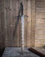 Hand forged Damascus fantasy sword with wave blade and barbed guard, standing upright against a wooden wall beside its leather scabbard