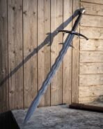 Hand forged Damascus fantasy sword with wave blade and blackened fittings, leaning at an angle against wooden boards on a stone ledge