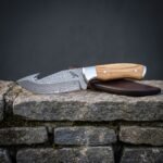 Hand forged Damascus gut hook knife with olive wood handle, engraved raven and brown leather sheath resting on a mossy limestone wall