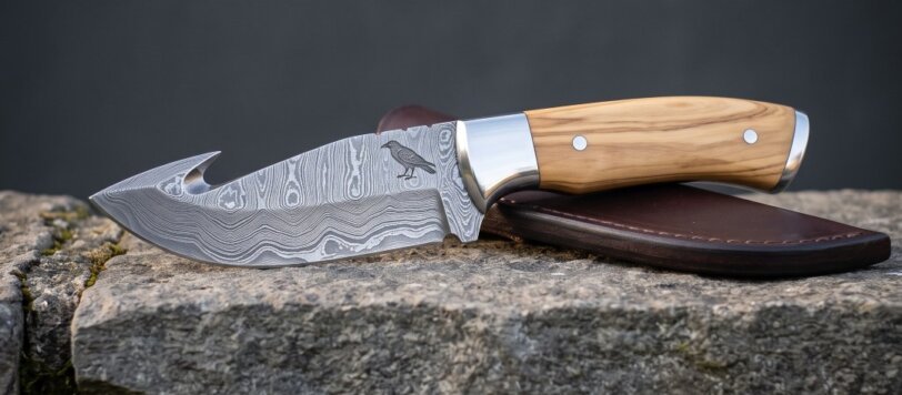 Hand forged Damascus gut hook knife with olive wood handle, engraved raven and brown leather sheath resting on a mossy limestone wall