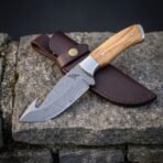 Hand forged Damascus gut hook skinning knife with olive wood handle angled across its brown leather belt sheath on stone