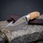 Hand forged Damascus gut hook skinning knife with olive wood handle, close view of the hook and patterned blade on stone
