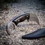 Hand forged Damascus karambit knife with ebony handle, close view of the finger ring and blade on gravel by a black sheath