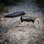 Hand forged Damascus karambit knife with ebony handle and finger ring standing on a gravel path with its black leather sheath behind