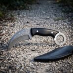 Hand forged Damascus karambit knife with ebony handle and steel finger ring, side view of the hooked blade on gravel