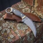 Hand forged Damascus karambit knife with micarta scales and steel finger ring resting on its brown leather sheath on lichen covered brick