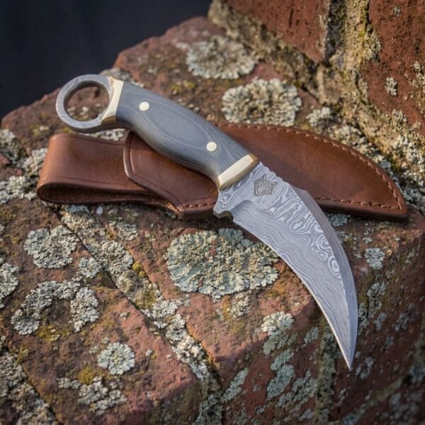 Hand forged Damascus karambit knife with micarta scales and steel finger ring resting on its brown leather sheath on lichen covered brick