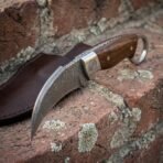 Hand forged Damascus karambit knife with rosewood scales, close view of the engraved bolster and curved blade lying on its leather sheath