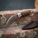 Hand forged Damascus karambit knife with rosewood scales and finger ring resting on lichen covered brick beside a dark leather sheath