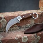 Hand forged Damascus karambit knife with rosewood scales and steel finger ring, side view over a brick step with its sheath