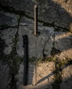 Hand forged Damascus katana with cord wrapped handle and oval iron tsuba, top down view of the hilt and black saya on flagstones