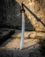 Hand forged Damascus katana with oval iron tsuba and curved blade, leaning against a sunlit stone wall with its saya lying behind