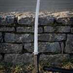 Hand forged damascus katana with a triskelion engraved above a brass habaki and a black cord wrapped tsuka standing against a dry stone wall beside its black saya