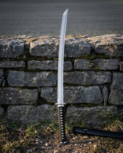 Hand forged damascus katana with a triskelion engraved above a brass habaki and a black cord wrapped tsuka standing against a dry stone wall beside its black saya