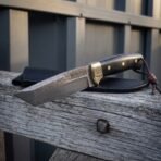 Hand forged Damascus knife with black scales and brass bolster resting on a weathered fence rail over its black sheath