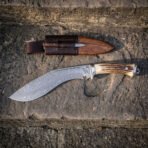 Hand forged Damascus kukri with antler handle, full side profile on a stone step below its leather sheath and companion blades