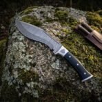 Hand forged Damascus kukri with black horn grip, overhead view of the curved blade on lichen stone beside the karda and chakmak sheath