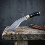 Hand forged Damascus kukri with black horn handle, side view at the edge of a stone bench against a dark slate wall
