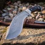 Hand forged Damascus kukri knife with black scales and steel bolster, close up of the patterned blade on stone with the sheath behind