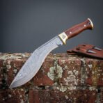 Hand forged Damascus kukri knife with brass bolster and rosewood handle, side view on a lichen covered brick wall with sheath