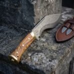 Hand forged Damascus kukri knife with olive wood scales and brass bolster, etched blade close up on a stone ledge
