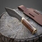 Hand forged Damascus kukri knife with rosewood handle, view from the brass pommel toward the blade on a stump with its sheath