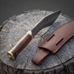 Hand forged Damascus kukri knife with rosewood handle and brass bolster, low side view on a cut stump by its belt sheath