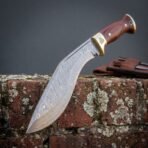 Hand forged Damascus kukri knife with rosewood scales, curved blade pattern shown leaning on a brick wall beside its sheath