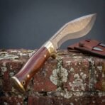Hand forged Damascus kukri knife with rosewood scales and brass bolster standing upright on an old brick wall by its sheath