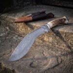 Hand forged Damascus kukri knife with stag antler handle and steel bolster, angled on a stone step with the leather sheath behind