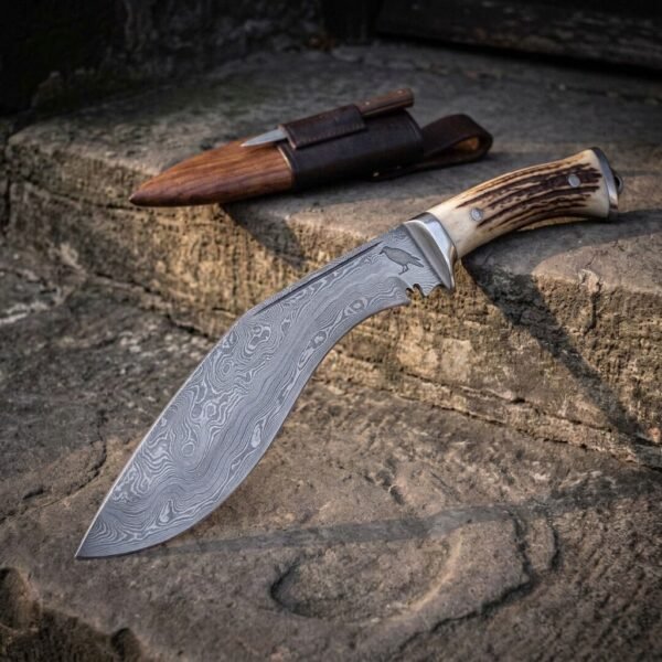 Hand forged Damascus kukri knife with stag antler handle and steel bolster, angled on a stone step with the leather sheath behind