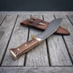 Hand forged Damascus kukri knife with walnut scales and brass bolster lying on weathered decking beside its leather sheath