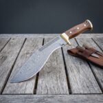 Hand forged damascus kukri knife with walnut scales and brass bolster resting on weathered grey decking beside a brown leather sheath holding two small companion knives