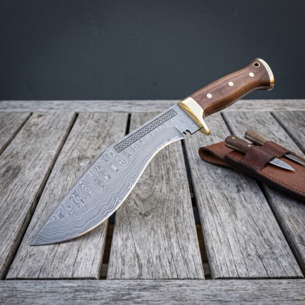 Hand forged damascus kukri knife with walnut scales and brass bolster resting on weathered grey decking beside a brown leather sheath holding two small companion knives