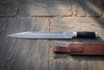 Hand forged Damascus seax knife with buffalo horn handle lying on paving stones with its brown leather belt sheath below the blade