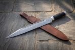 Hand forged Damascus seax knife with buffalo horn handle laid diagonally across its brown leather belt sheath on grey flagstones