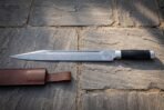 Hand forged Damascus seax knife with buffalo horn handle, full length side view on paving with the leather sheath at left