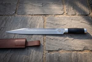 Hand forged Damascus seax knife with a wolf head etch and dark buffalo horn handle lying on stone flagstones beside a brown leather belt sheath in warm light