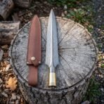Hand forged damascus spearhead with a brass socket collar and leaf shaped blade beside a brown leather sheath on a weathered tree stump in woodland.