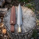 Hand forged damascus spearhead with a brass socket collar and leaf shaped blade beside a brown leather sheath on a weathered tree stump in woodland.