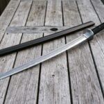 Hand forged Damascus steel black saya katana with a cord wrapped handle and round steel tsuba resting beside its dark stained wooden scabbard on weathered grey decking