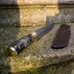 Hand forged Damascus steel broken back seax with horn handle and brass fittings, on a mossy stone step next to its dark sheath