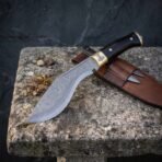 Hand forged Damascus steel kukri knife with black horn handle and brass bolster, on a weathered stone bench beside its sheath