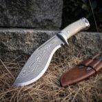 Hand forged Damascus steel kukri knife with bone handle scales, patterned blade on dry grass by a stone ledge with its sheath