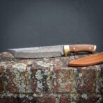 Hand forged Damascus tanto blade and walnut grip shown side on along a brick wall, tan leather sheath set apart