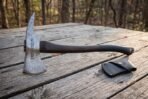 Hand forged Damascus tomahawk with a bearded head, upswept horn and Elder Futhark runes on a dark walnut haft resting on a weathered picnic table in woodland