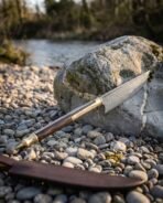 Hand forged Damascus Viking bowie knife with rosewood handle leaning on a riverside boulder over pebbles, leather sheath in front