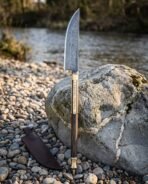 Hand forged Damascus Viking bowie knife with rosewood handle standing upright against a mossy rock on a pebble bank