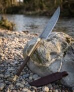 Hand forged Damascus Viking bowie knife with rosewood handle and brass bolster propped point up on a riverside rock