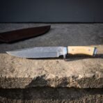 Hand forged damascus viking bowie knife with pale bone handle scales and a Valknut etched blade resting on rough stone beside its oxblood leather belt sheath.